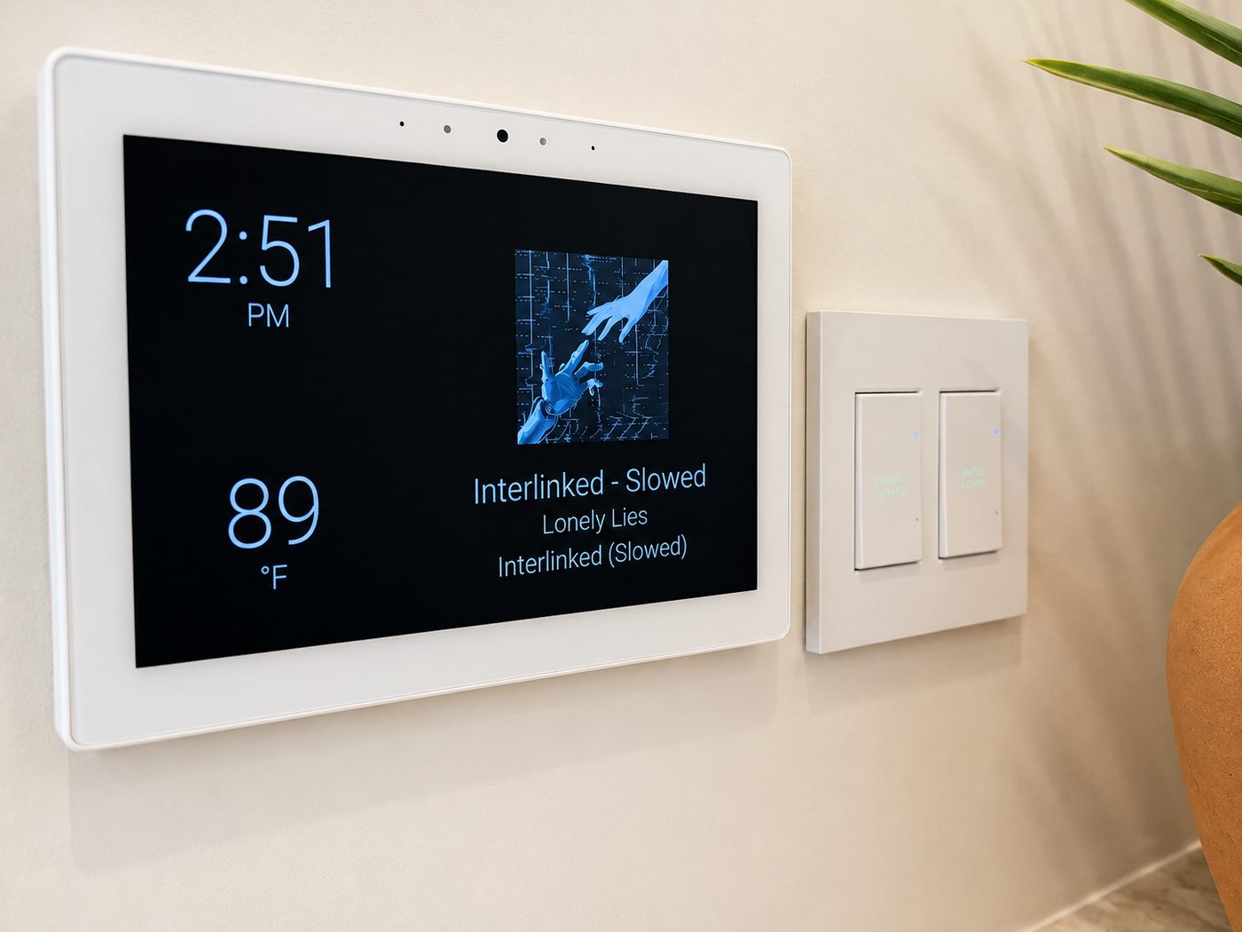 Wall-mounted smart home touchscreen showing room temperature and now-playing audio next to a lighting keypad